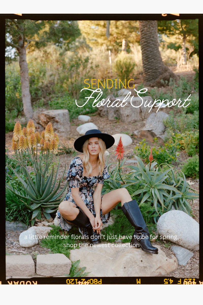 A woman wearing a floral dress, black wide-brim hat, and black knee-high boots sits outdoors in a garden setting with rocks and plants. The image has a vintage film-strip border and text overlays reading 'Sending Floral Support' and 'a little reminder florals don't just have to be for spring.