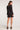 Full body back view of woman wearing the Saltwater Luxe Verra Mini Dress in black polka dot with long sleeves and black sandals on a white backdrop