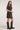 Full body side view of woman wearing the Saltwater Luxe Kove Mini Dress in Black with patterned details and black boots on a white background