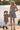 A woman and a young girl wearing matching strapless gingham mini dresses stand hand in hand by a lake. The woman wears a white cowboy hat and tall beige boots, while the girl wears dark boots and white socks