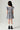 A young girl viewed from behind wearing a blue and white checkered mini dress with ruffled short sleeves, white socks, and black boots, standing against a light gray studio background