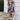 A young girl with pigtails stands outdoors against a white house wall wearing a dark floral short-sleeve dress with a ruffled neckline, white ruffled socks, and black ankle boots