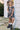 A young girl with pigtails stands outdoors against a white house wall wearing a dark floral short-sleeve dress with a ruffled neckline, white ruffled socks, and black ankle boots