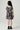 A young girl viewed from behind wearing a short-sleeve tiered dress with a dark navy floral print, white socks, and black boots against a light gray studio background