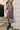 A young girl with pigtails stands outdoors against a white house wall wearing a short-sleeve ruffled dress with a brown and cream floral pattern, white ruffled socks, and black boots