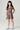 A young girl with long dark hair stands in a studio wearing a short-sleeve tiered dress with a brown, red, and white floral pattern, paired with white high-top sneakers