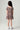 A young girl viewed from behind wearing a short-sleeve, smocked, tiered dress with a dark red and beige floral pattern, paired with white sneakers, against a light gray studio background