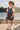 A young girl wearing a navy blue and white vertically striped pinafore dress over a white t-shirt, styled with white high-top sneakers and sunglasses, standing outdoors near a lake