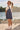 A young girl with a ponytail and sunglasses stands outdoors by a lake, wearing a navy blue and white vertically striped pinafore dress over a white t-shirt, white socks, and white high-top sneakers