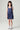 A young girl with dark hair stands against a light gray studio background wearing a sleeveless tiered dress with vertical blue and white stripes, and white sneakers