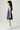 A young girl with long dark hair is shown in profile wearing a sleeveless tiered dress with vertical blue and white stripes, paired with white high-top sneakers, against a plain light gray studio background