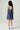 A young girl viewed from behind wearing a sleeveless blue and purple vertically striped pinafore dress with white sneakers, standing against a light gray studio background
